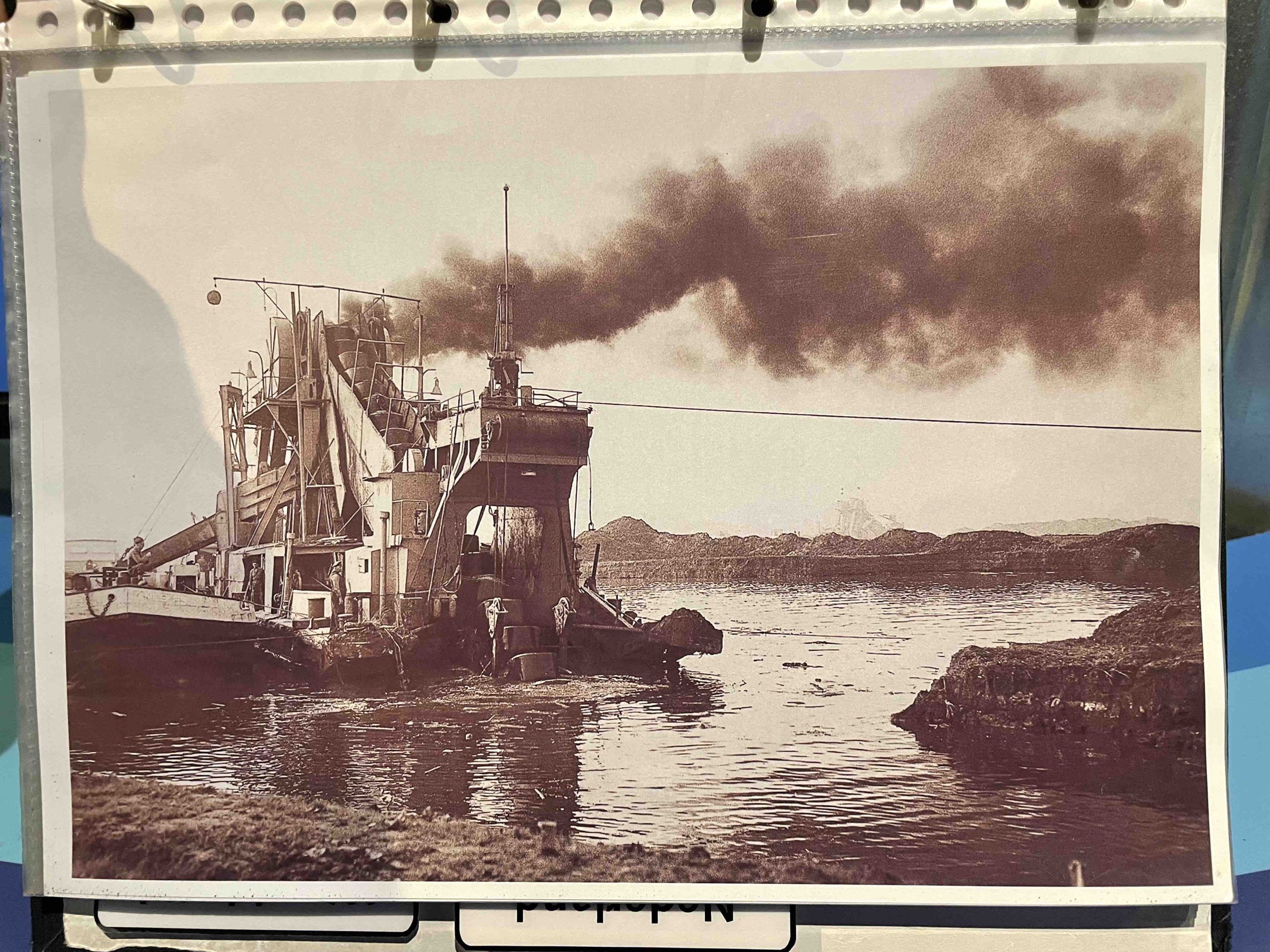 Mechanization of dredging. The dark plume of pollution in this historical photo also demonstrates the dependence on fossil energy to scale and increase the capacity, strenght, and effeciency of dredge capital. Machines also took over labour intensive tasks. Photo by Henrik Ernstson of collections at Publikszentrum Water, Rotterdam, May 2022.