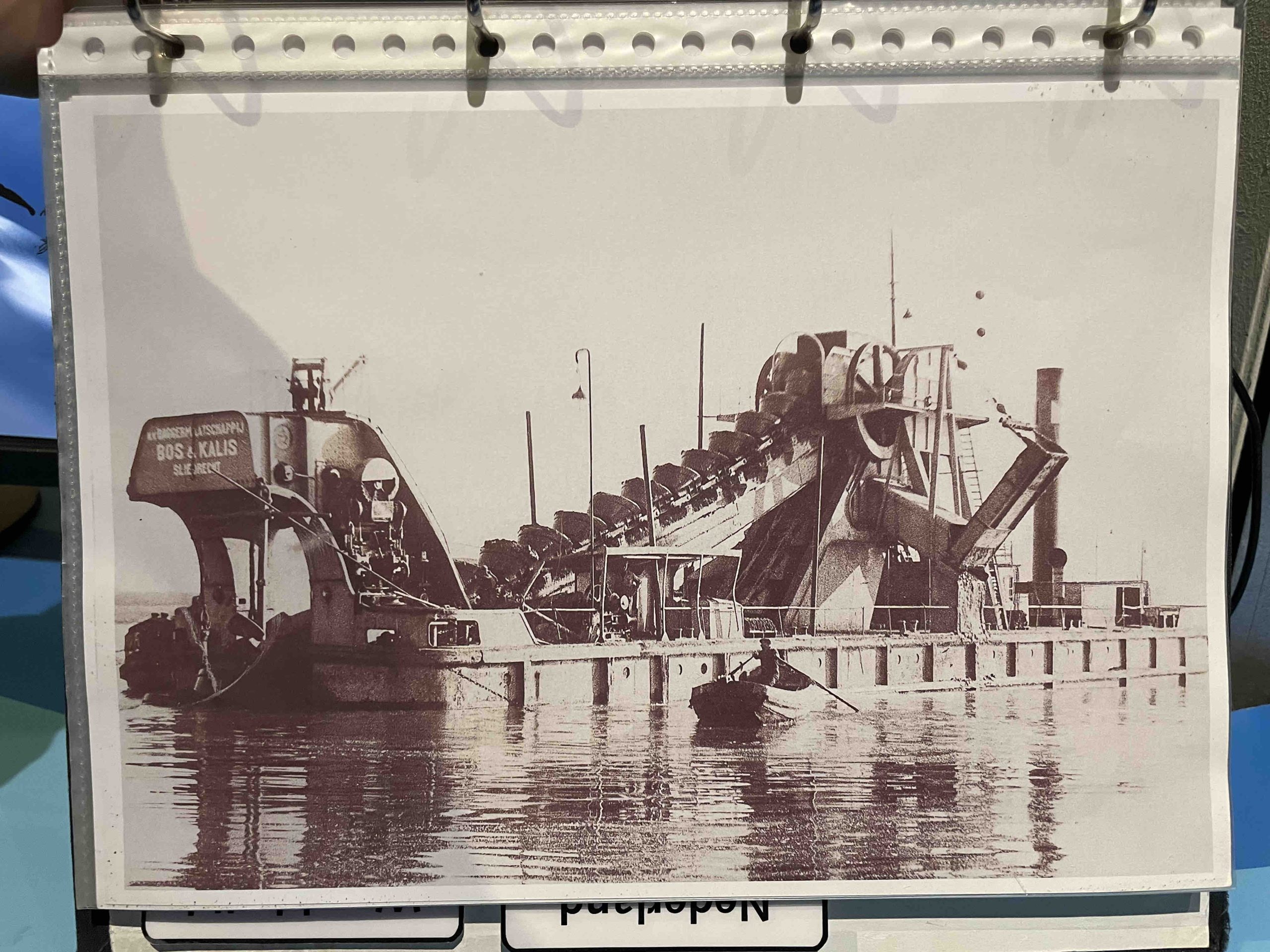 A bucket dredger from the company BOS & KALIS, today Boskalis, lying idle in the bay with a rowing worker approaching. Photo by Henrik Ernstson of collections at Publikszentrum Water, Rotterdam, May 2022.